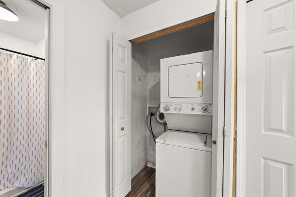 A laundry room with a stacked washer and dryer, a door with a doorknob, and a patterned shower curtain.
