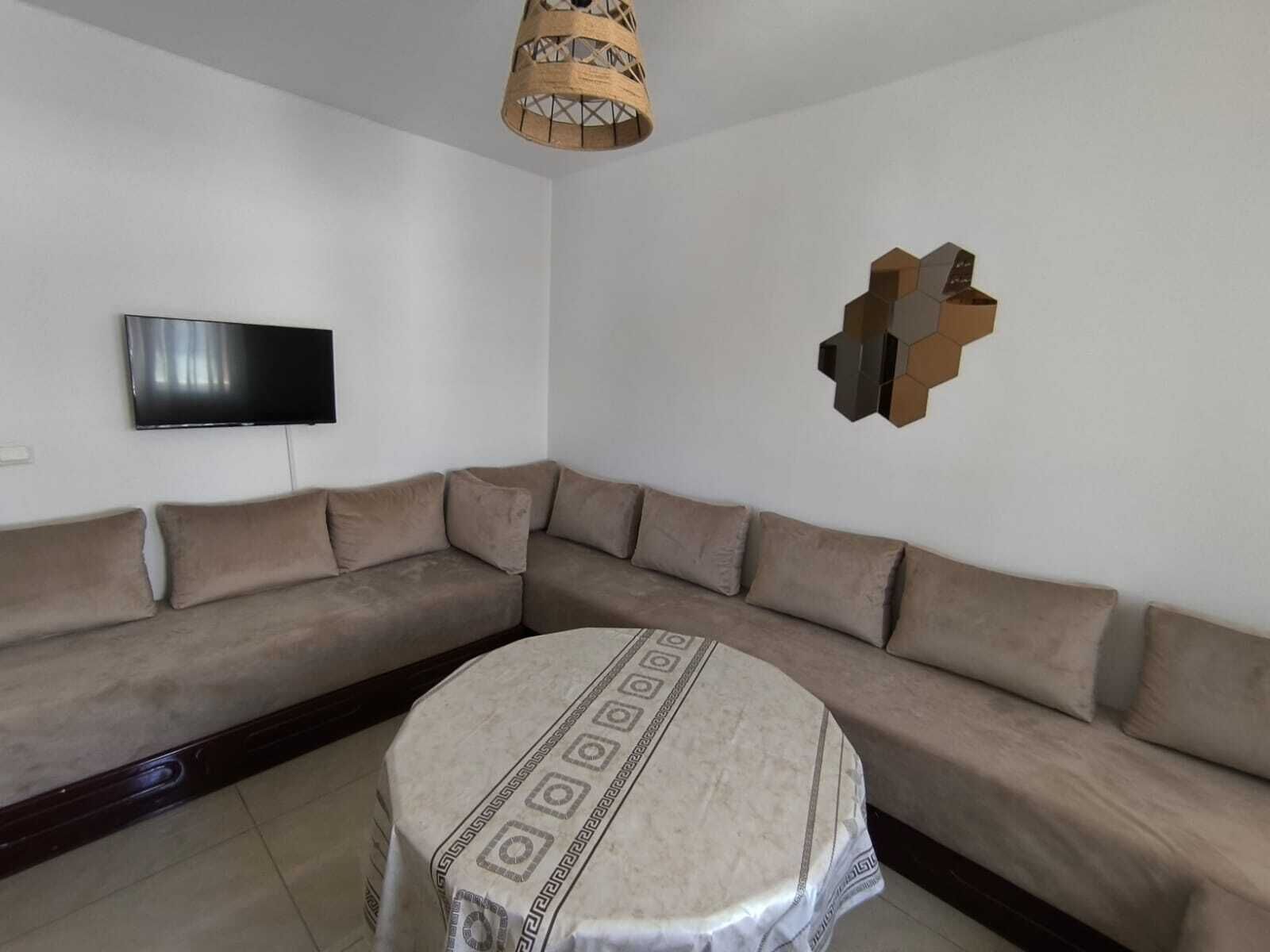 Un salon comprenant un canapé d’angle beige, une table basse ronde recouverte d’une nappe à motifs, un téléviseur à écran plat fixé au mur et un élément décoratif géométrique mural.