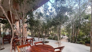 An outdoor dining area with wooden tables and chairs, surrounded by trees and a stone wall. - Hermosa Villa en Tulum (Tulum)