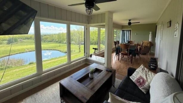 Un vaste salon doté de grandes fenêtres donnant sur un lac, un coin repas avec des chaises et une table, et un ventilateur de plafond.