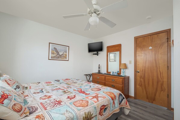 A bedroom with a bed, a ceiling fan, a television, a dresser, a mirror, and a door.