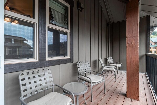 A covered porch with four metal chairs and a small round table, wooden floor, and a window with a view of a house.