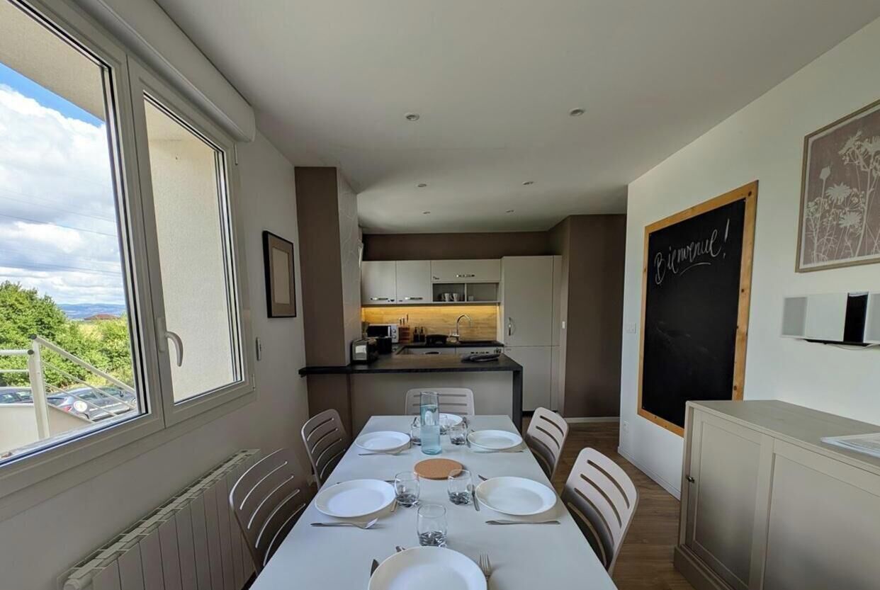 A dining area with a long table set for four, a kitchen in the background, and a chalkboard with 'Bonjour!' written on it.