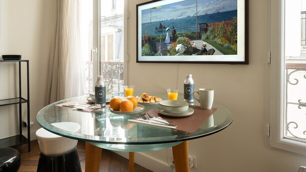 Une table de petit-déjeuner avec un plateau en verre, un tableau encadré représentant un paysage côtier, et une vue sur la mer par la fenêtre.