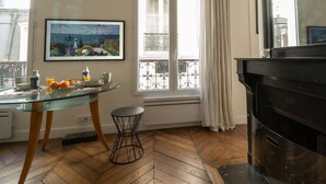 Un espace repas avec une table en verre, un parquet en bois disposé en chevrons, un piano noir et un balcon doté d’une balustrade.