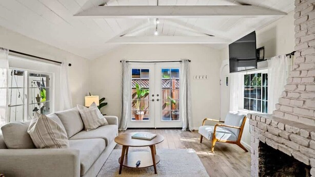A spacious living room with a high ceiling, a fireplace, a sofa, a coffee table, and a view through a glass door to an outdoor area with plants. - Carmel Cozy Cottage | Room for 4 (Carmel)