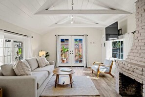 A spacious living room with a high ceiling, a fireplace, a sofa, a coffee table, and a view through a glass door to an outdoor area with plants. - Carmel Cozy Cottage | Room for 4 (Carmel)