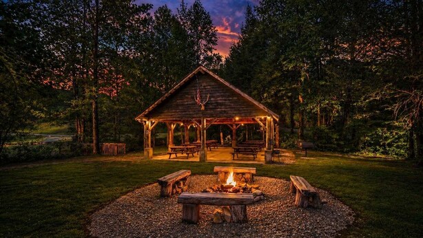 A wooden gazebo with benches and a fire pit in a wooded area.