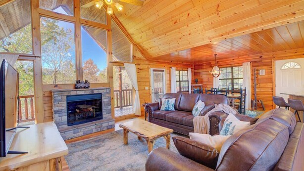 A cozy cabin interior with a stone fireplace, leather sofa, wooden dining table, and large windows offering views of the outdoors. - 4BR Pigeon Forge Cabin | Hot Tub, Sauna, Game Room | 1 Mi to Dollywood, Pool Access, Scenic Views (Pigeon Forge)