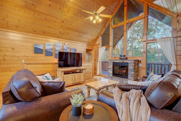 Interior acogedor de una cabaña con chimenea, sofás de cuero, televisor y ventanas amplias.