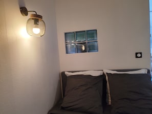 A bed with two pillows, a wall-mounted light fixture, and a window with blinds. - Charming 25m2 studio close to CDG airport, Parc Astérix, sandy sea, (Louvres)