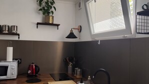 A compact kitchen with a microwave, kettle, and toaster. A window with blinds is on the right, and a plant is on the wall. - Charming 25m2 studio close to CDG airport, Parc Astérix, sandy sea, (Louvres)
