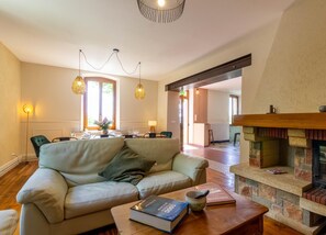 Een woonkamer met een bank, een open haard en een eethoek op de achtergrond.