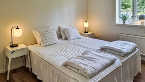 Een net opgemaakt tweepersoonsbed met een gewatteerde deken, twee nachtkastjes met lampen, en een raam met uitzicht op groen buiten.