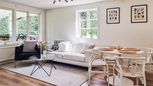 Een lichte, ruim verlichte kamer met een witte bank, een zwarte fauteuil, een glazen salontafel en een witte eettafel met geweven placemats. Er zijn twee ramen met planten op de vensterbank, en ingelijste botanische prints aan de muur.