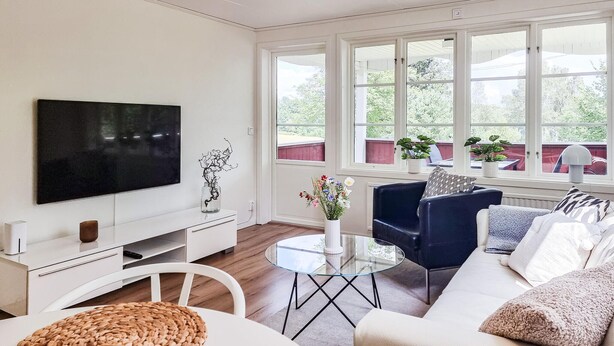 Een moderne woonkamer met een flatscreen televisie, een glazen salontafel, een blauwe bank en een witte zithoek. Op de vensterbank staan planten en op de vloer ligt een geweven vloerkleed.