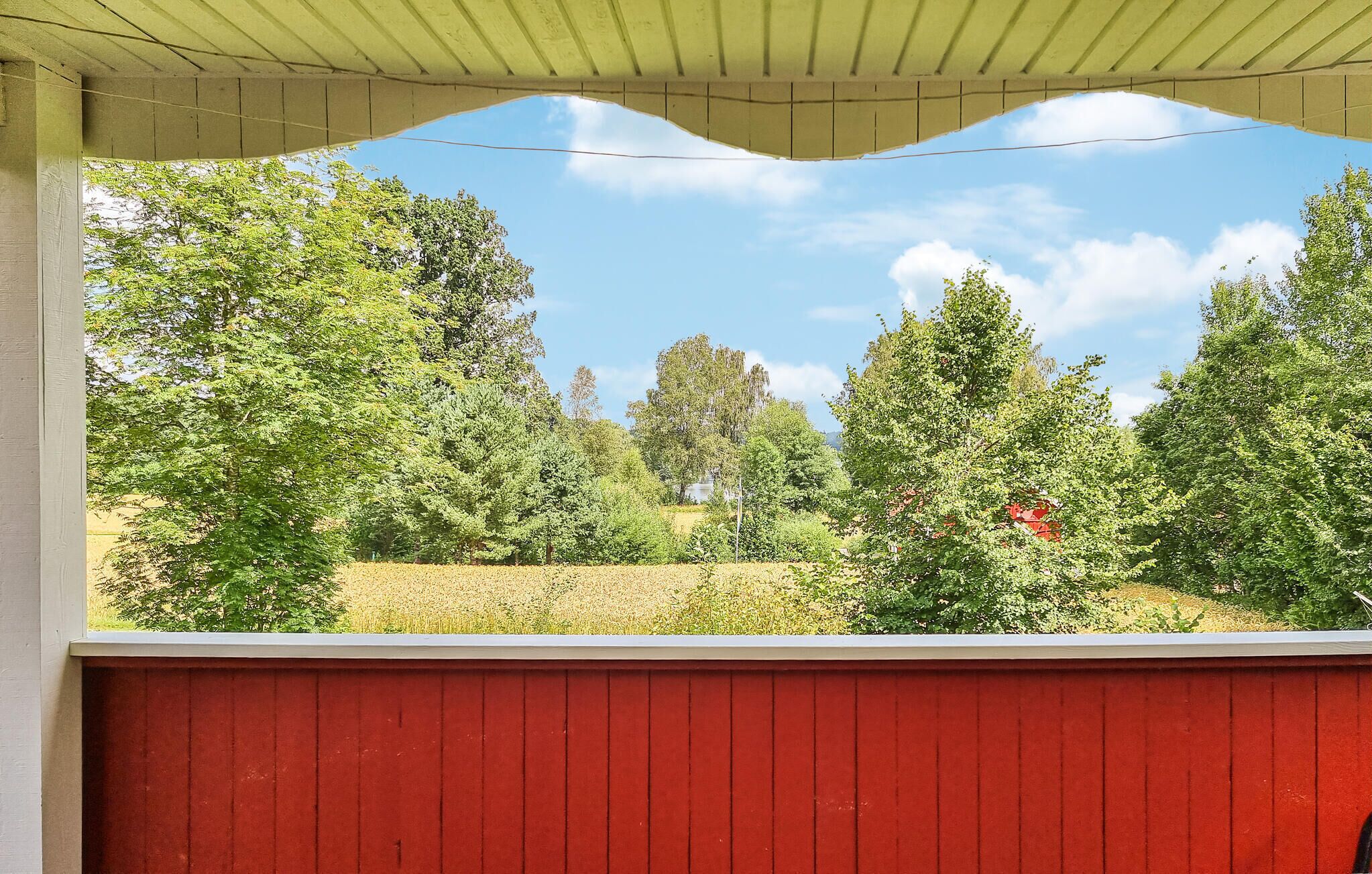 Eine überdachte Veranda mit roter Geländer, die auf einen Garten mit verschiedenen Bäumen und blauem Himmel blickt.