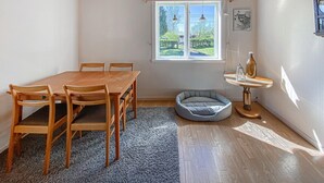 Un comedor con mesa y sillas de madera, una cama para mascotas, una pequeña mesa redonda con una botella y un vaso, y una ventana con vistas a zonas verdes.