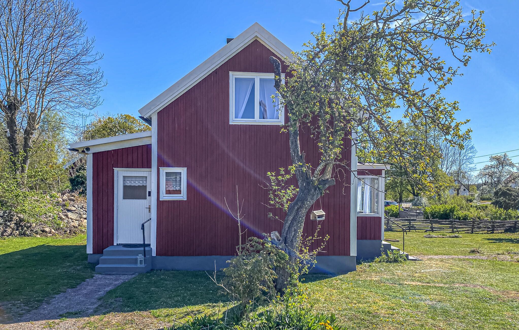 A red wooden house with a white door and windows, surrounded by greenery and a clear sky.