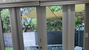 Una habitación con puerta corrediza de vidrio que da a un jardín con piscina, mobiliario exterior y un patio cubierto.