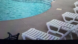 A swimming pool with a water fountain, surrounded by lounge chairs and a covered area with a building in the background.
