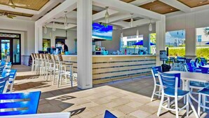 A covered outdoor seating area with white and blue chairs, a counter with a person behind it, and a TV screen displaying a blue sky.