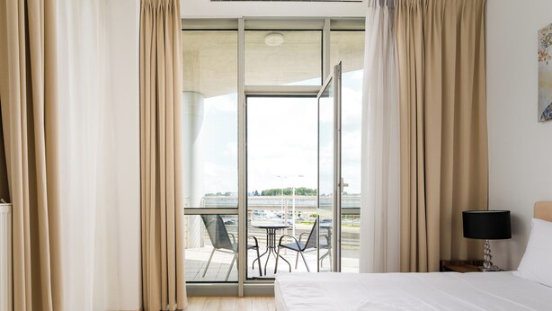 Een hotelkamer met een bed, nachtkastje, lamp, raam met gordijnen en balkon met tafel en stoelen.