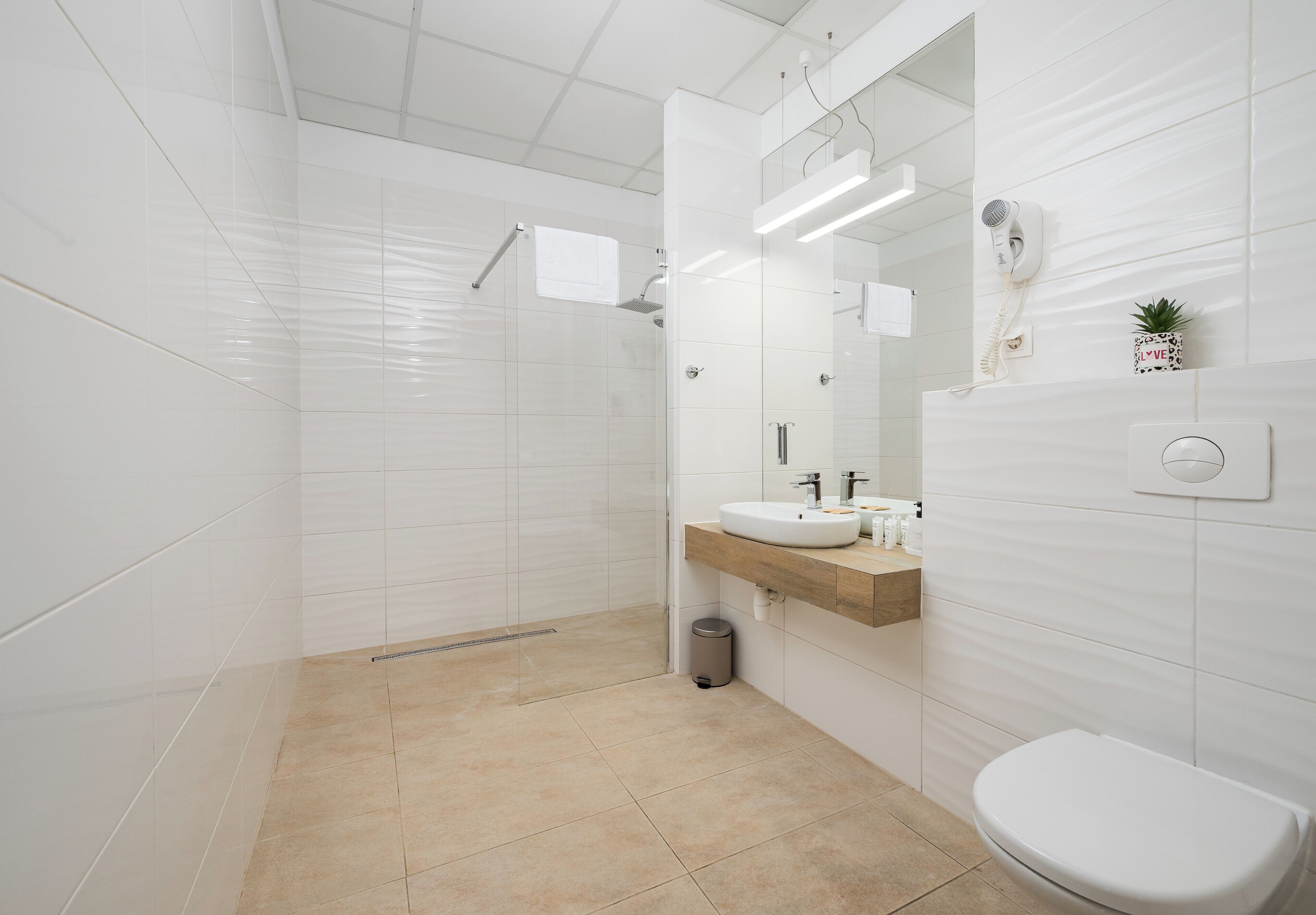 Ein modernes Badezimmer mit weißen Fliesen, einem beigen Boden, einem Holzwaschtisch, einem weißen Waschbecken, einem Spiegel, einem Föhn und einer kleinen Pflanze.