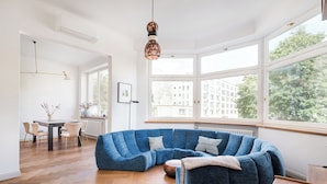 A modern living room with a large blue sectional sofa, wooden herringbone flooring, a dining table with chairs, and large windows offering a view of buildings and trees. - Mokotowska Superior Apartment (Warszawa)