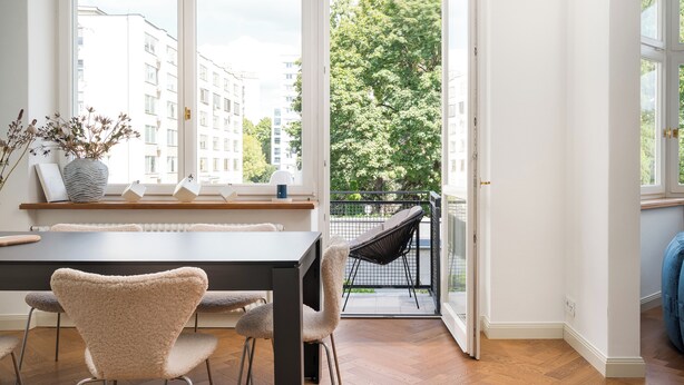 A modern dining area with a table and chairs, large windows with a view of buildings and trees, and a balcony with a chair. - Mokotowska Superior Apartment (Warszawa)