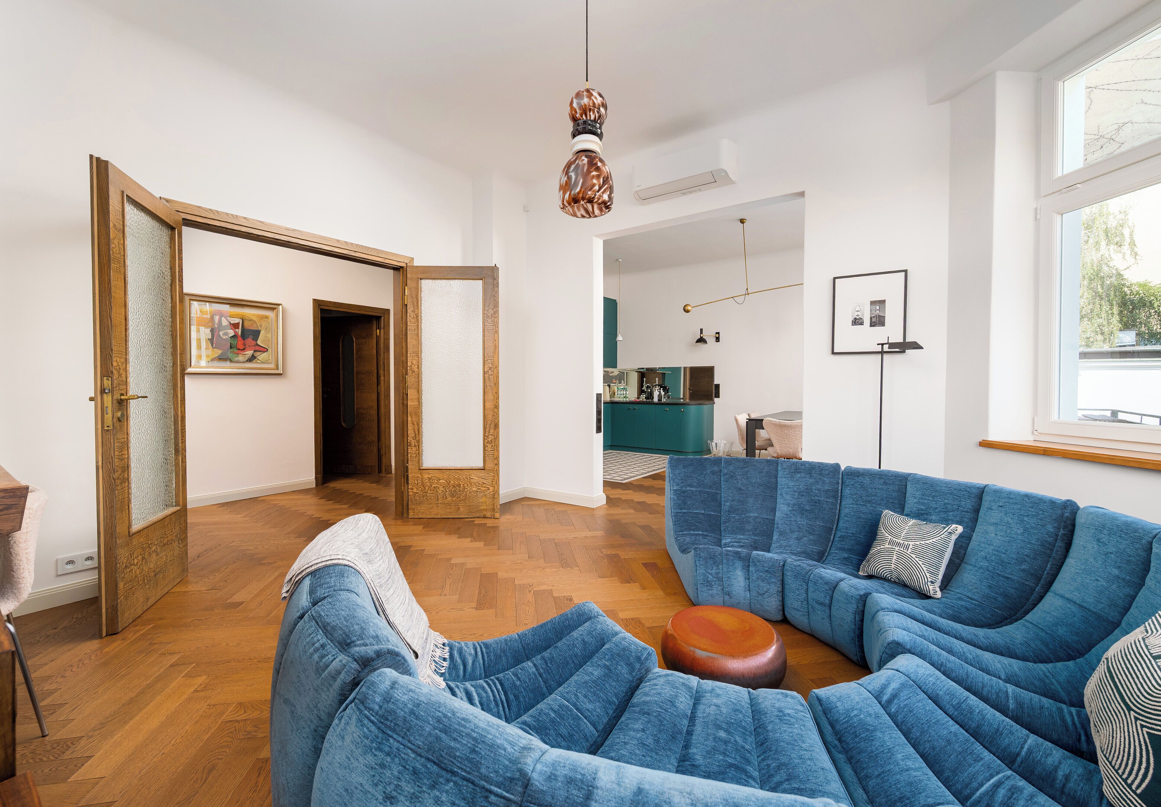Een moderne woonkamer met een blauwe zithoek, een houten vloer en een keuken op de achtergrond.