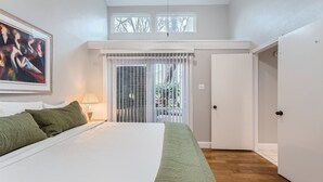 A bedroom with a large bed, a green throw blanket, a painting on the wall, and a window with blinds.