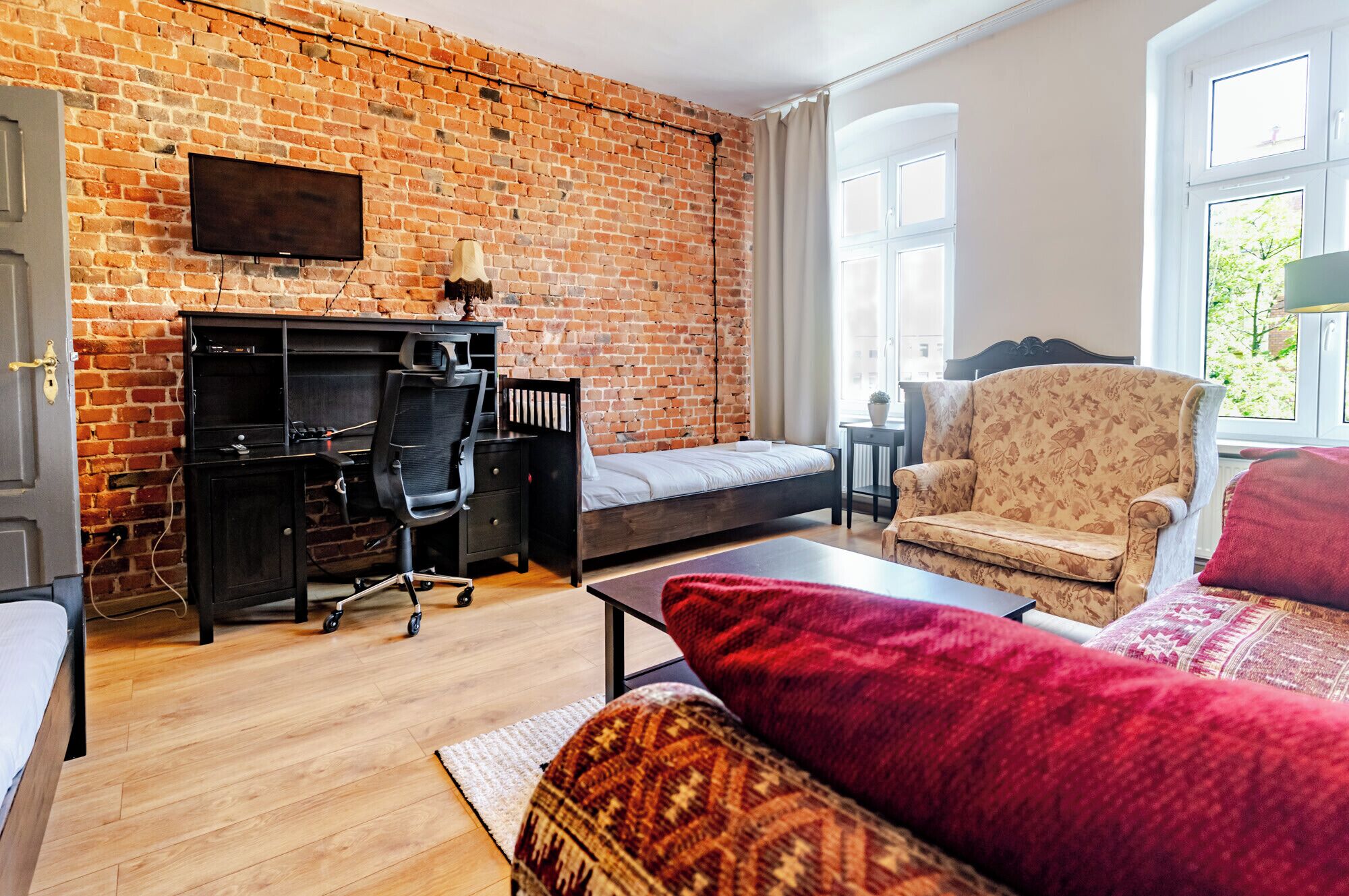 A room with a brick wall, a bed, a desk with a chair, a sofa, and a window with a view of greenery.