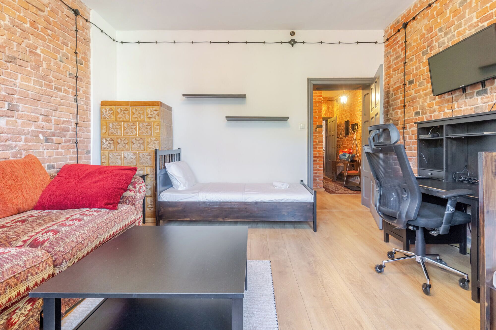 A compact living space with a bed, a sofa, a small table, a TV mounted on the wall, and a fireplace.