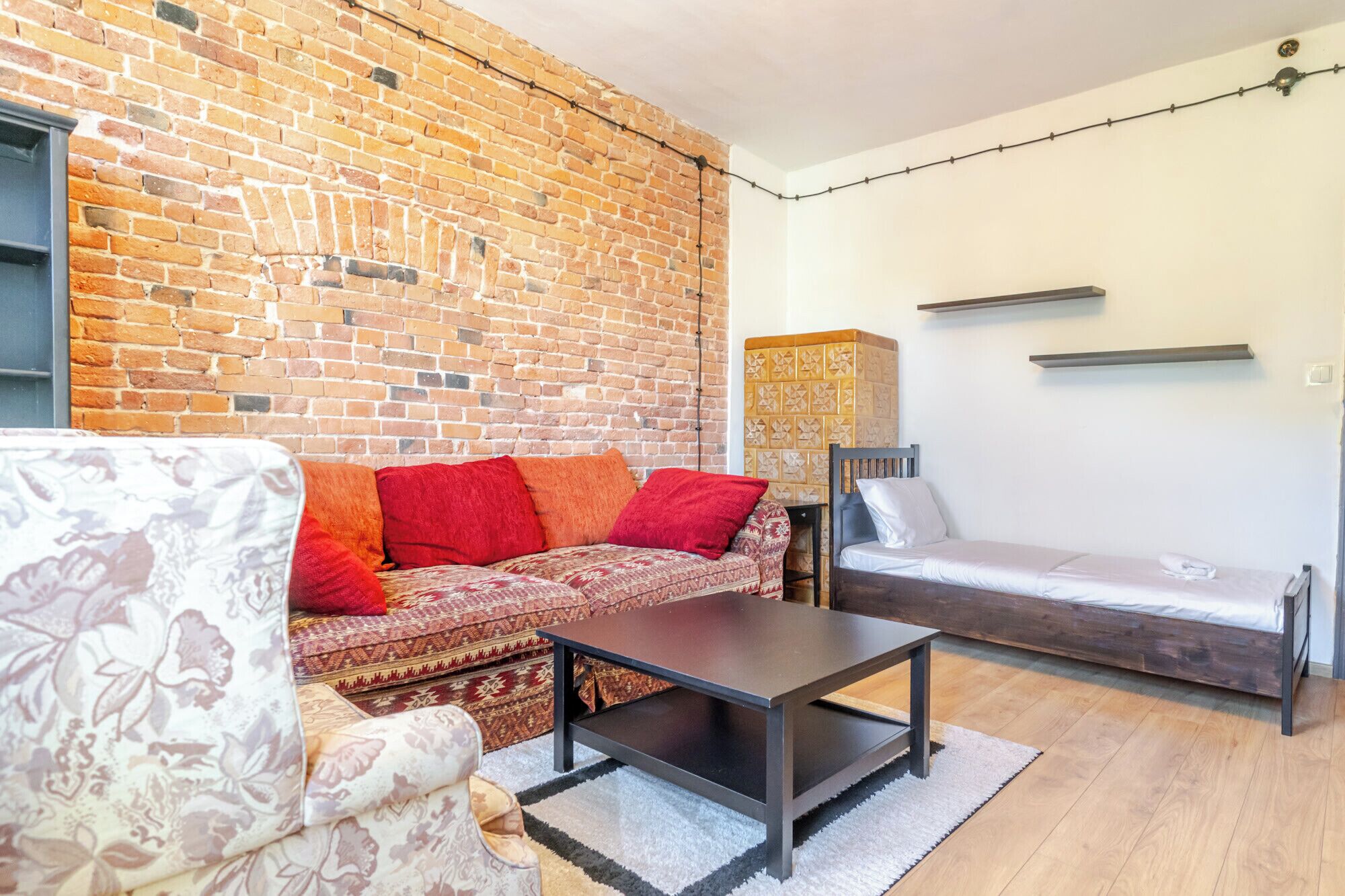 Ein gemütliches Wohnzimmer mit Backsteinwand, einem gemusterten Sofa, einem kleinen Tisch und einem Bett mit weißer Matratze.
