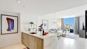 A modern kitchen with a marble island, a dining area with a white sofa, and a view of the cityscape through large windows. - FTL Tiffany Topaz: 7th FL, Rooftop Pool, Gym, Walk to Beach (Fort Lauderdale)