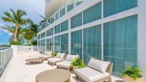 A modern outdoor seating area with a round wooden table and cushioned chairs, large windows, and palm trees in the background. - FTL Tiffany Topaz: 7th FL, Rooftop Pool, Gym, Walk to Beach (Fort Lauderdale)