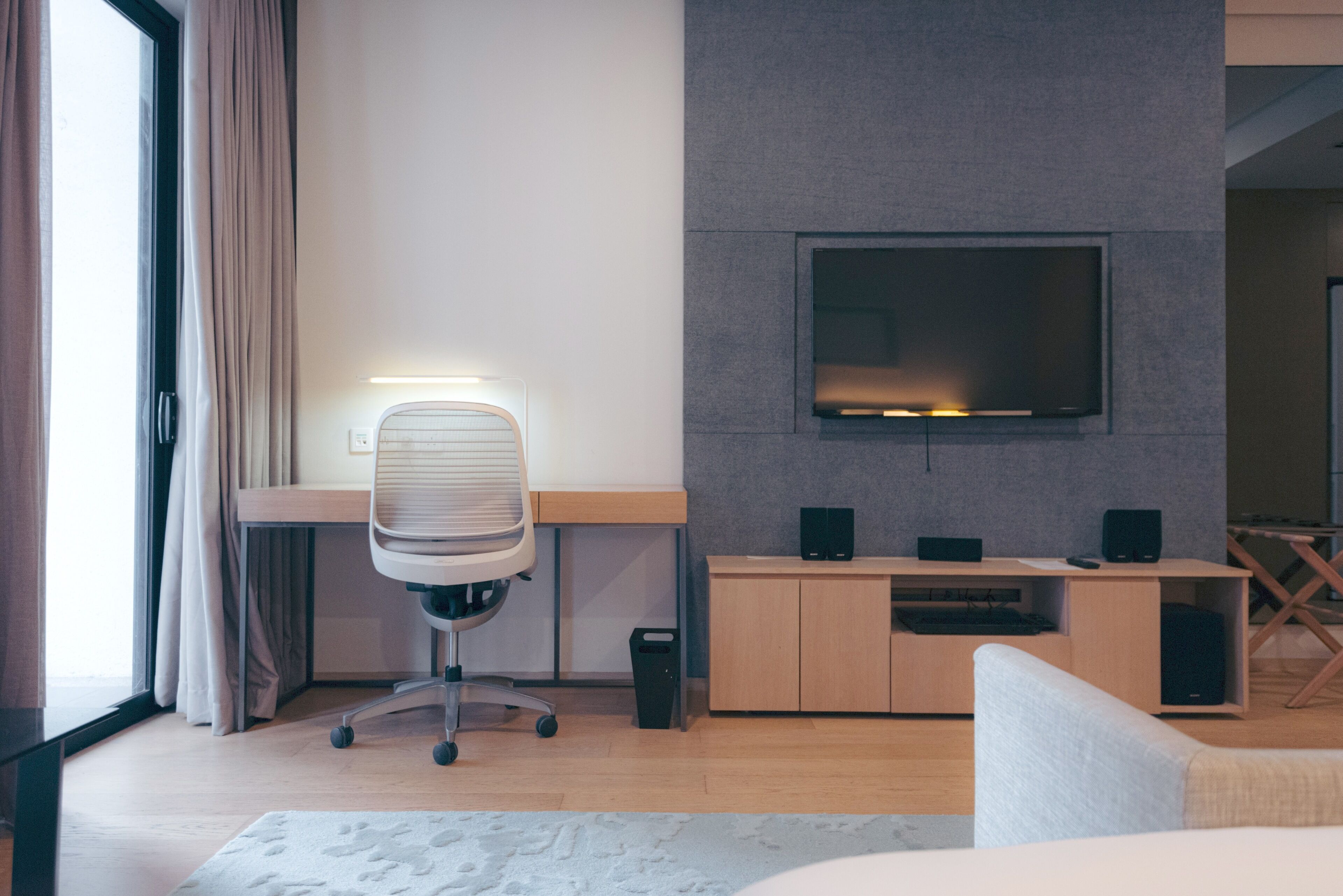 Une chambre d’hôtel moderne dotée d’un bureau en bois, d’un fauteuil confortable, d’un téléviseur à écran plat fixé au mur, d’un petit meuble contenant des haut-parleurs et d’une grande fenêtre avec des rideaux.