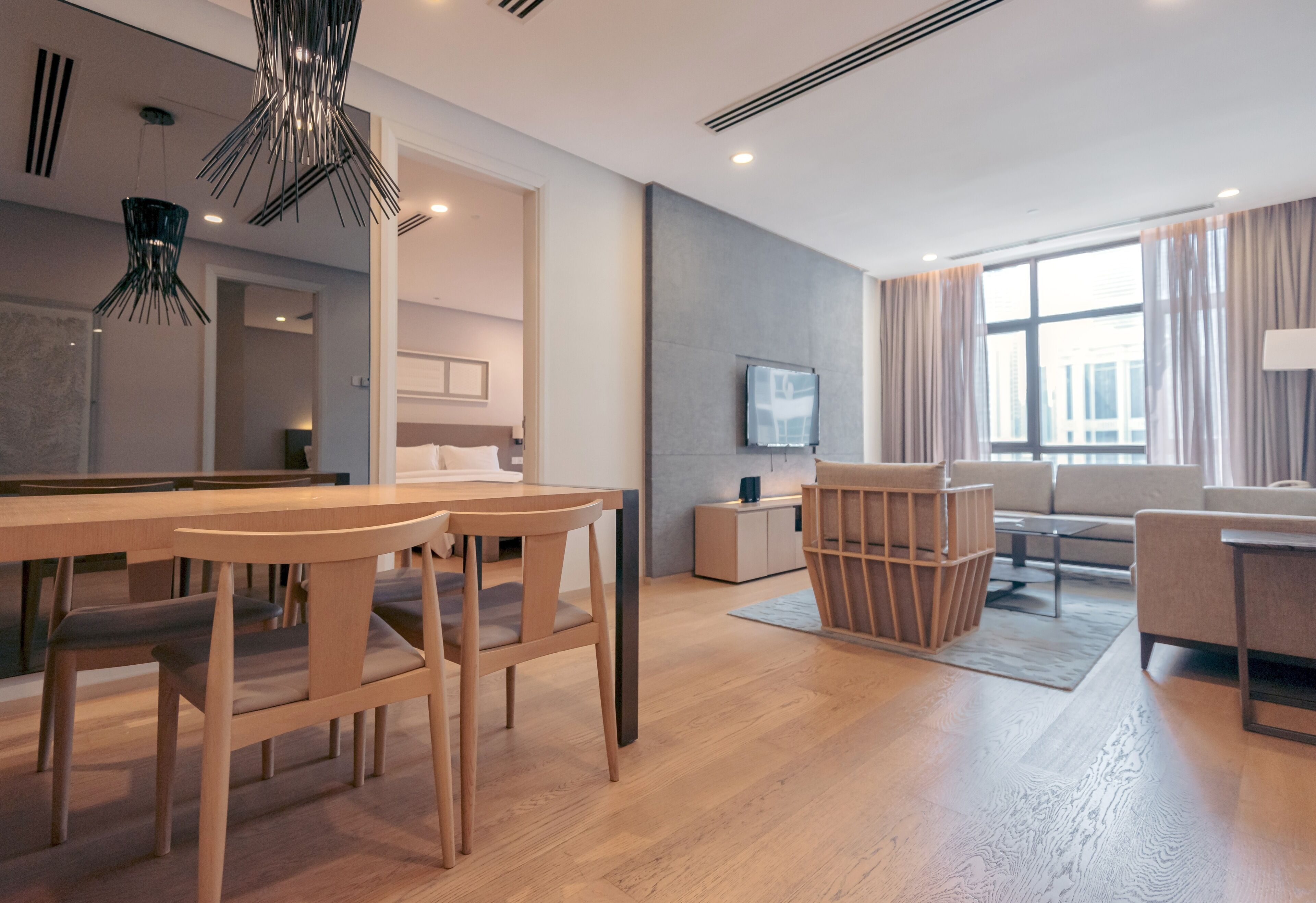 A modern hotel room with a wooden dining table, chairs, a sofa, and a TV mounted on the wall.