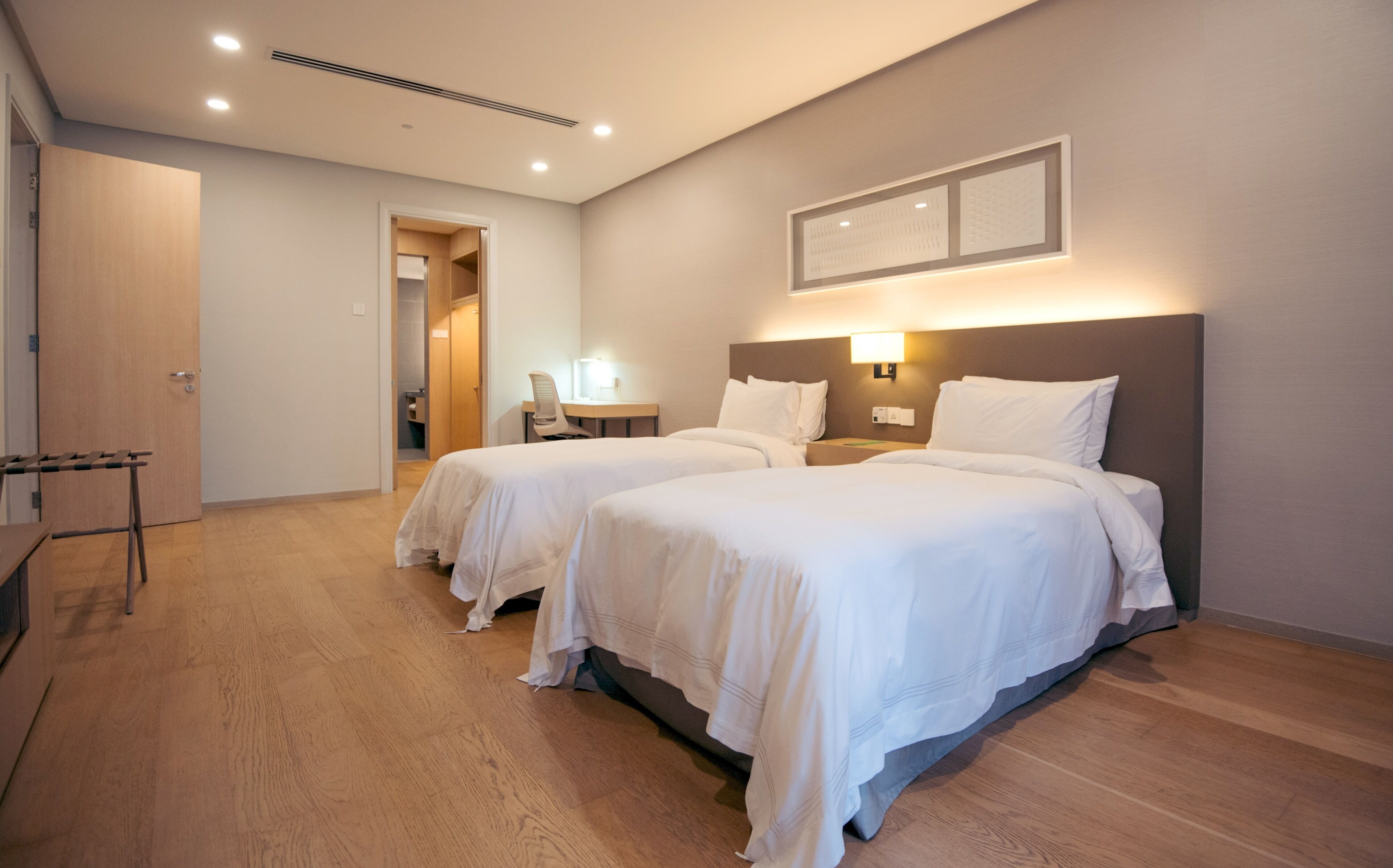 Quarto de hotel com duas camas, piso de madeira, uma escrivaninha com cadeira e luminária fixada na parede.