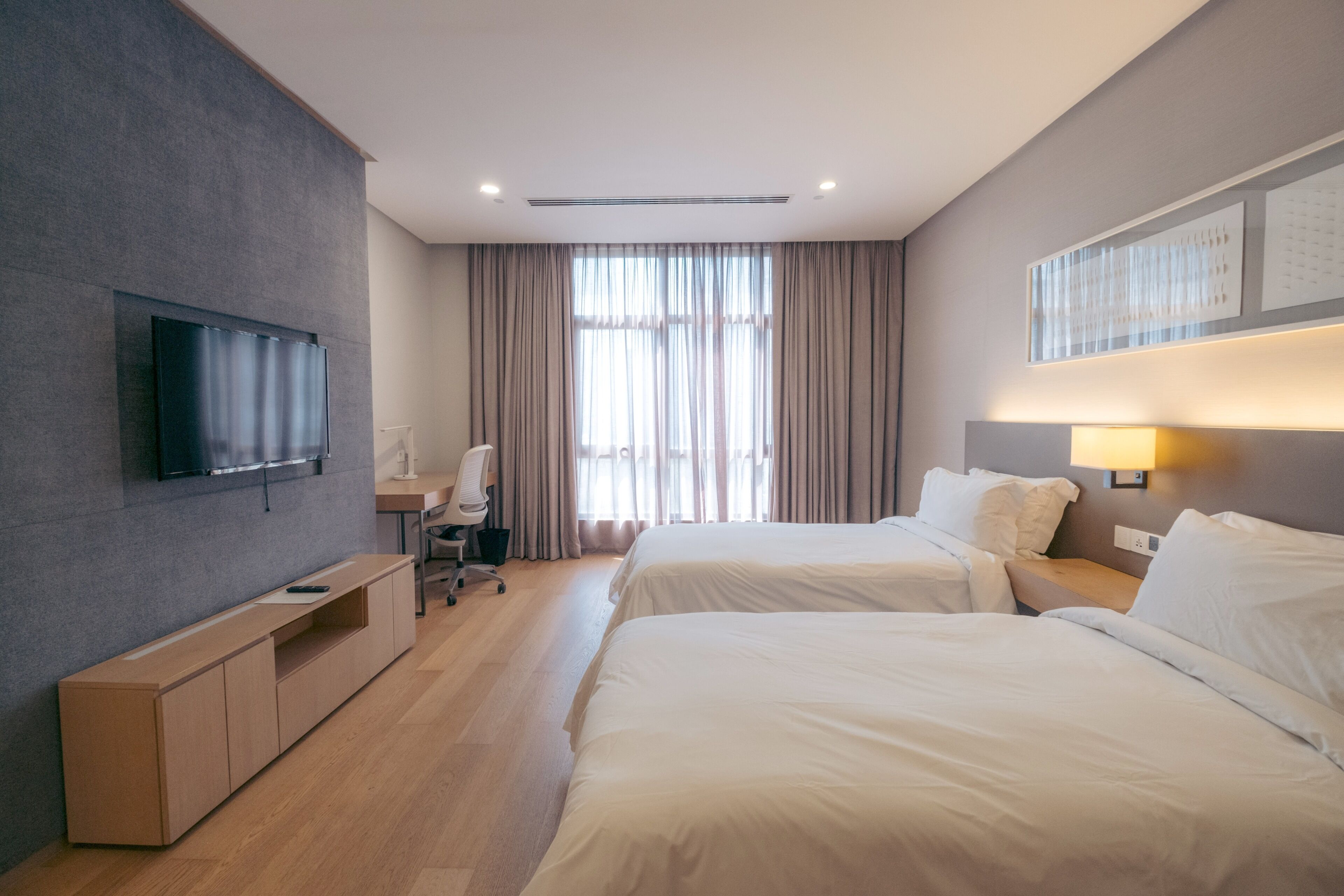 A hotel room with two beds, a TV mounted on the wall, a desk with a chair, and a large window with curtains.
