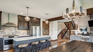 A modern kitchen with a large island, bar stools, and a wooden staircase. - Hawk Ridge in the Highlands of Breck | Golf Course and Nordic Trails! (Breckenridge)