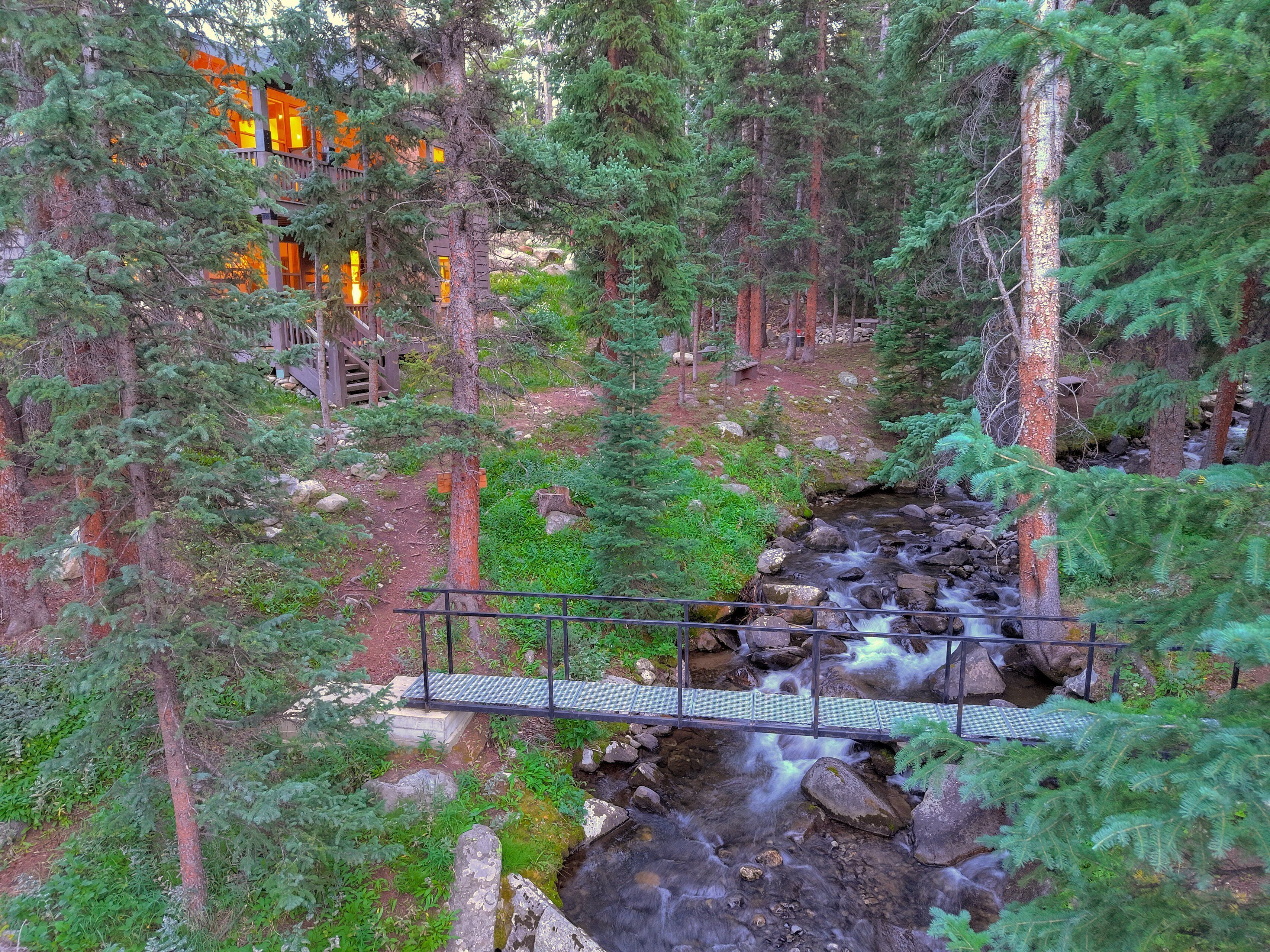 A forested area with a small bridge over a stream, surrounded by trees and a building with orange windows.