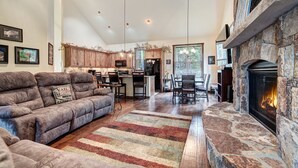 A spacious living room with a stone fireplace, a large sofa, and a dining area with a kitchen in the background.