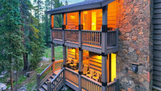 A multi-level wooden cabin with a stone exterior, featuring a balcony with chairs and a staircase leading to the upper level.