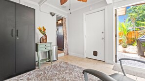 A room with a large wardrobe, a small desk with a vase, a ceiling fan, and a view of a garden through an open door. - Cozy 2BR Snowbird Home Near Wilton Manors (Fort Lauderdale)