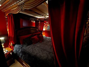 Una habitación con cama, cortinas rojas y una mesita de noche con lámpara.