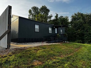 A modern, dark-colored building with a flat roof, a small porch with chairs, and a gravel pathway leading to the entrance. - Sunset Cabin-Adults Only Hot Tub Getaway (Hillsboro)