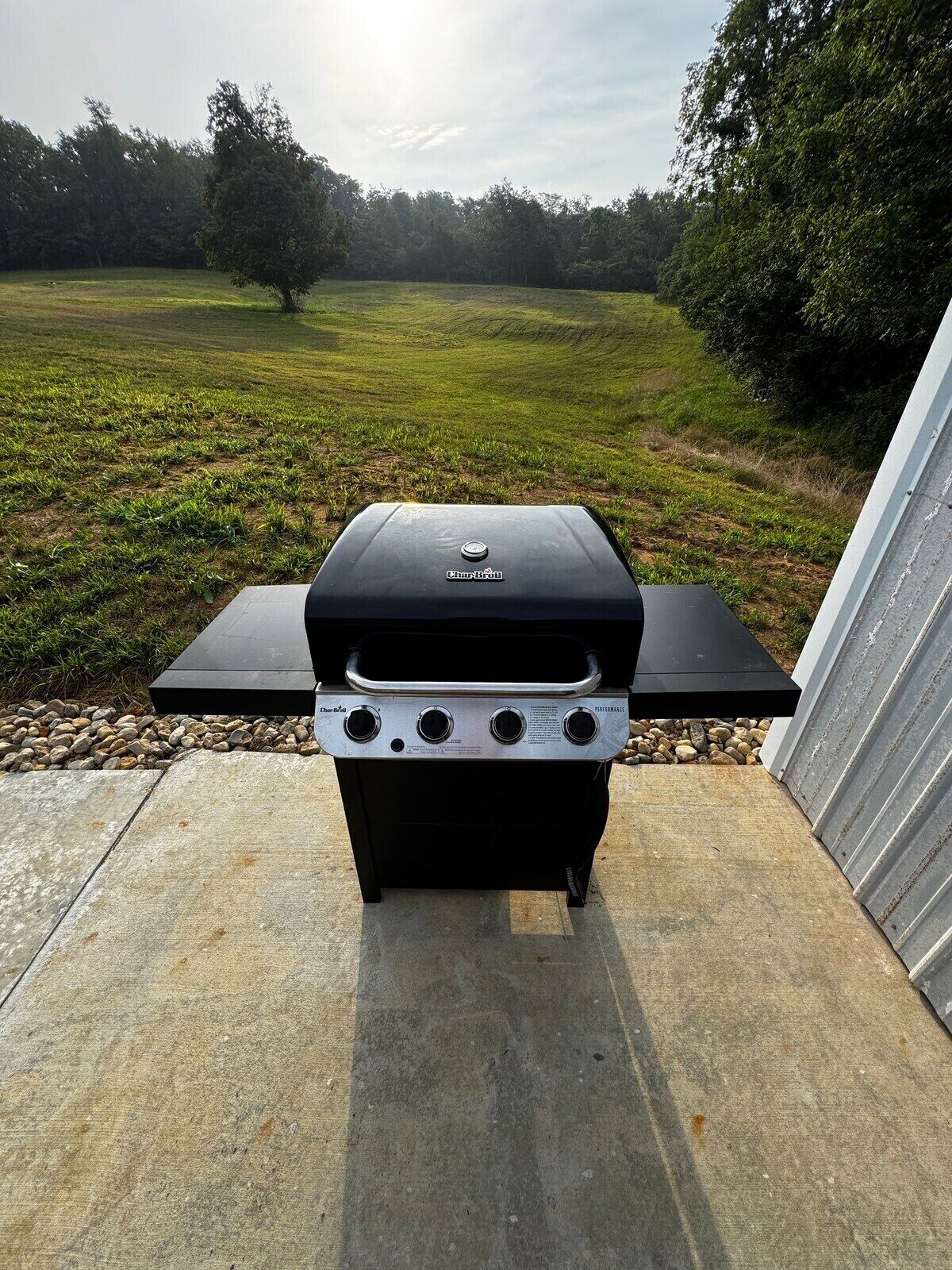 Una parrilla de gas Char-Broil con tapa negra y tres perillas de control, ubicada en un patio de concreto con un campo de césped y árboles al fondo.