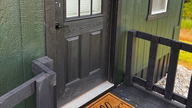 A dark green wooden building with a black door, a window, and a small balcony with a 'hello' mat. - Sunset Cabin-Adults Only Hot Tub Getaway (Hillsboro)